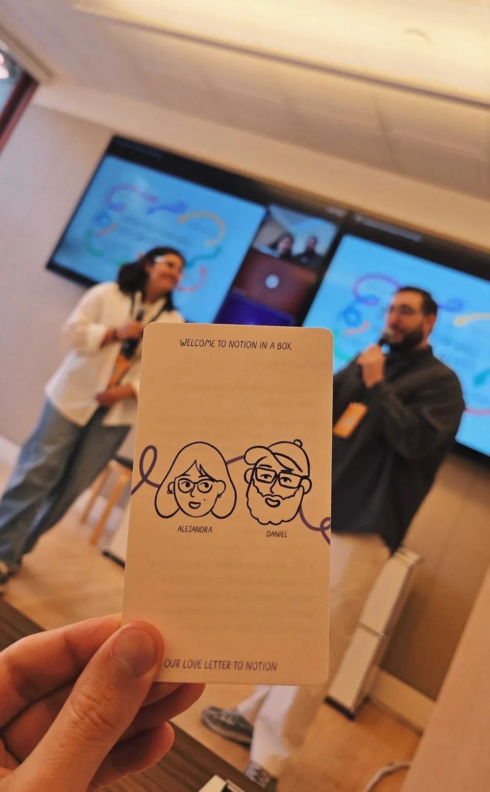 A hand holding up the welcome card, with drawings of Alejandra and Daniel, at the Notion Ambassadors Summit while two presenters speak behind it.